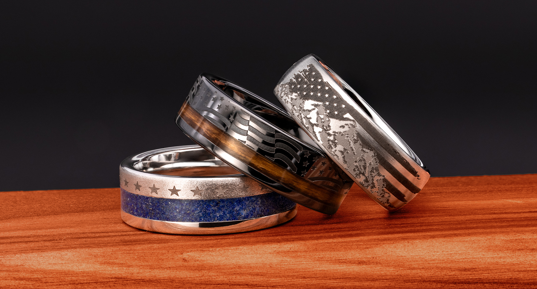 Photo of three mens rings featuring blue lapis inlay, stars engravings, flag engravings, mountains with flag engraving, and bourbon barrel wood inlay sitting on red wood.